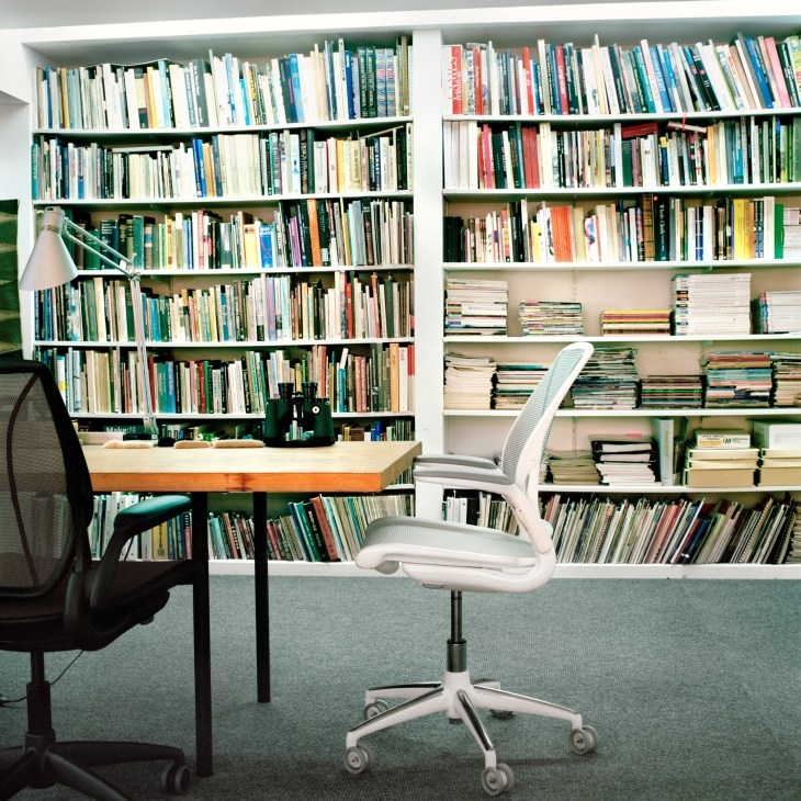 A home library with floor-to-ceiling white shelving filled with books, a wooden desk, and ergonomic office chairs in the foreground.