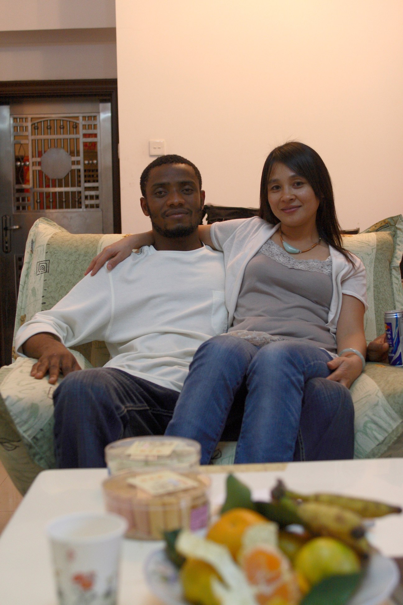 An African man and Chinese woman sitting together on a couch in a home interior.