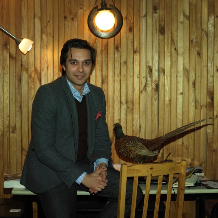 Correspondent Neave Barker sits on a wooden chair against a wood-paneled wall beneath a hanging lamp, with a taxidermied bird beside him.