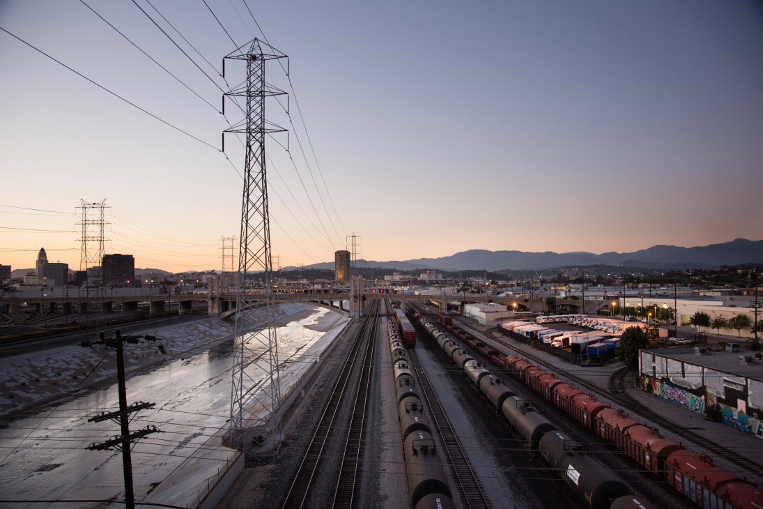 Los Angeles River and train yards with cargo trains, power lines, and downtown skyline at dusk.