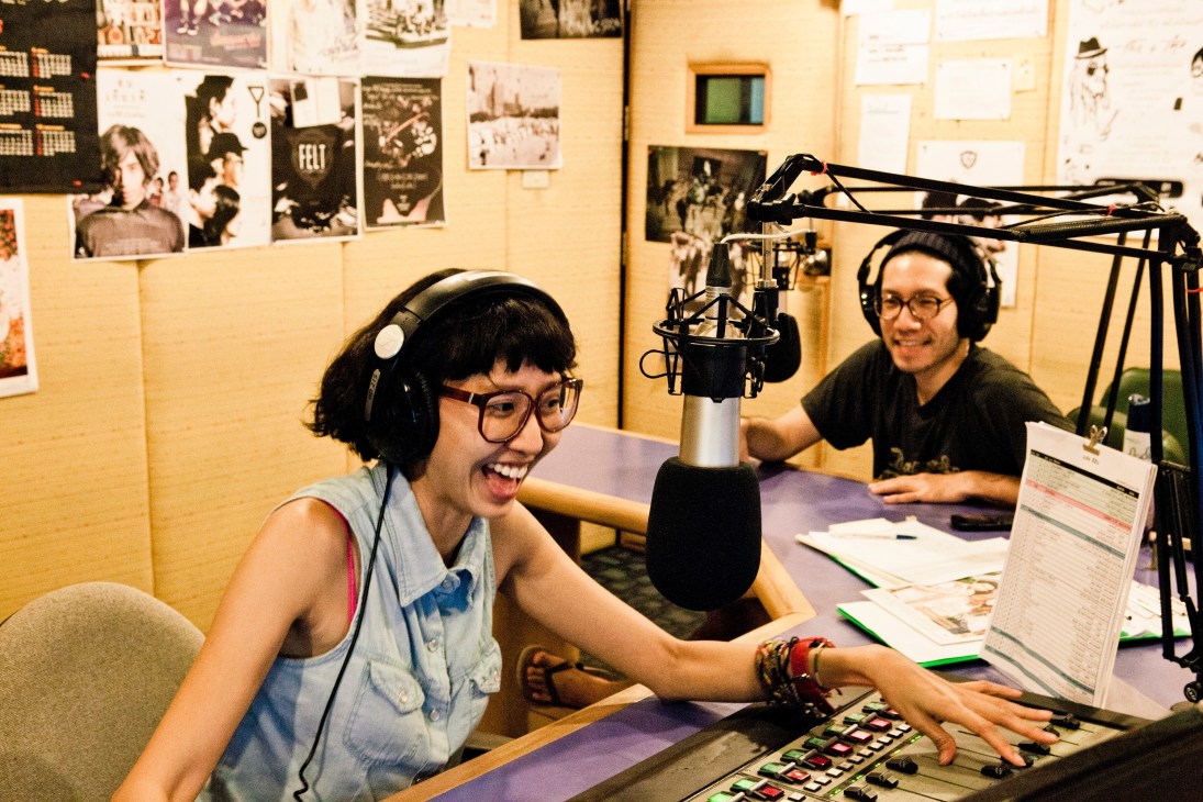 Two radio DJs at a broadcast station desk with microphone and equipment.