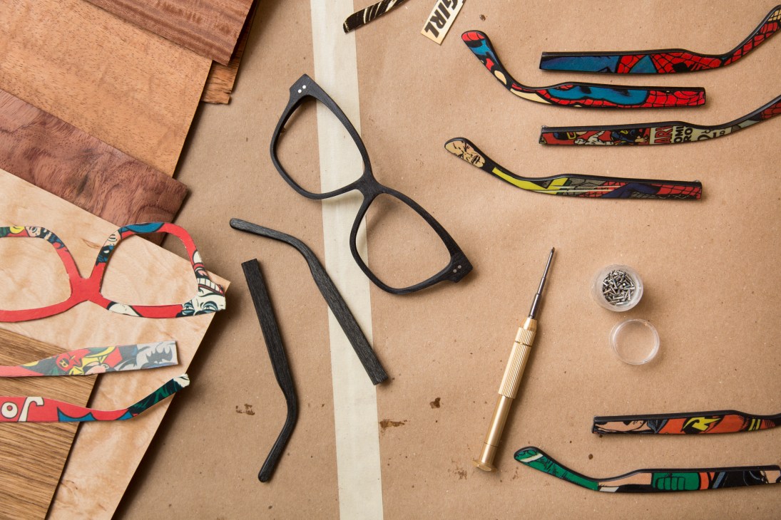Eyeglass frames in black and wood with comic-book patterned temple arms, reclaimed wood samples, and tools arranged on a kraft paper surface.