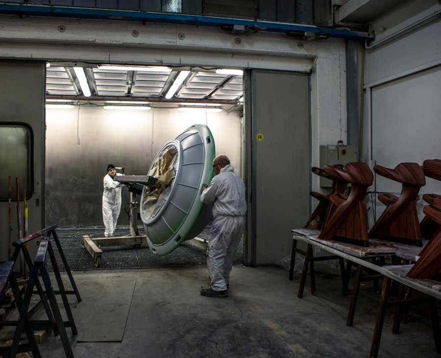Two workers in white protective suits assemble a large green and silver carousel swing ride component in an industrial factory workshop.