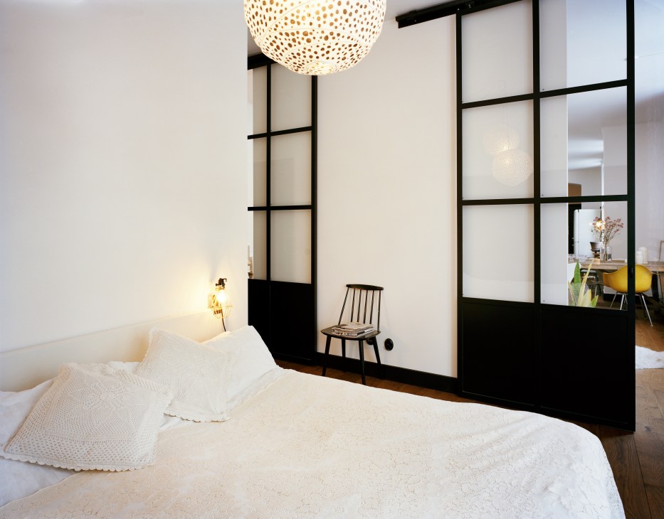 Modern bedroom with white bedding, black-framed glass partition doors, and a woven pendant light fixture.