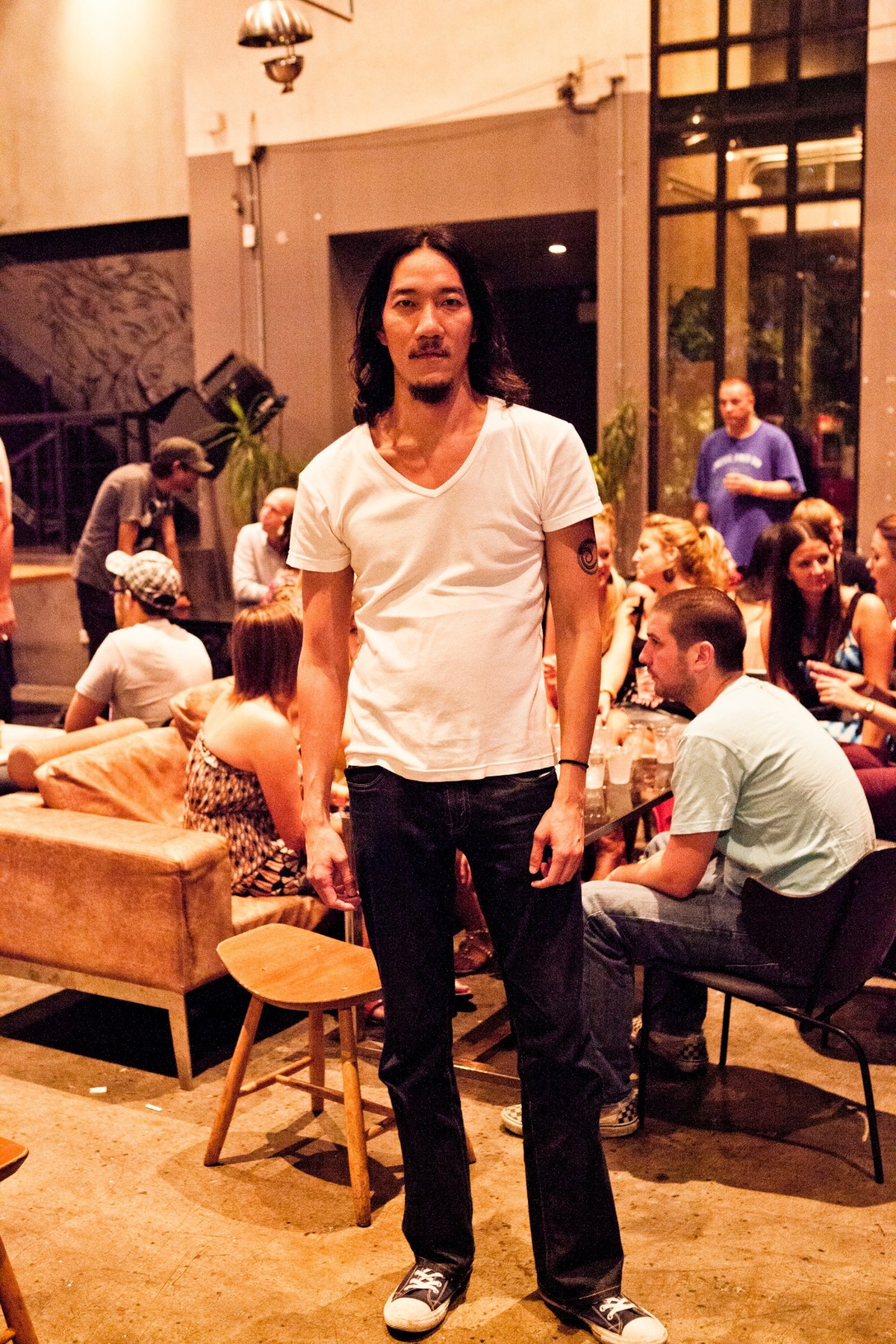 Man with long dark hair and handlebar mustache standing in a casual indoor venue with guests seated around him.