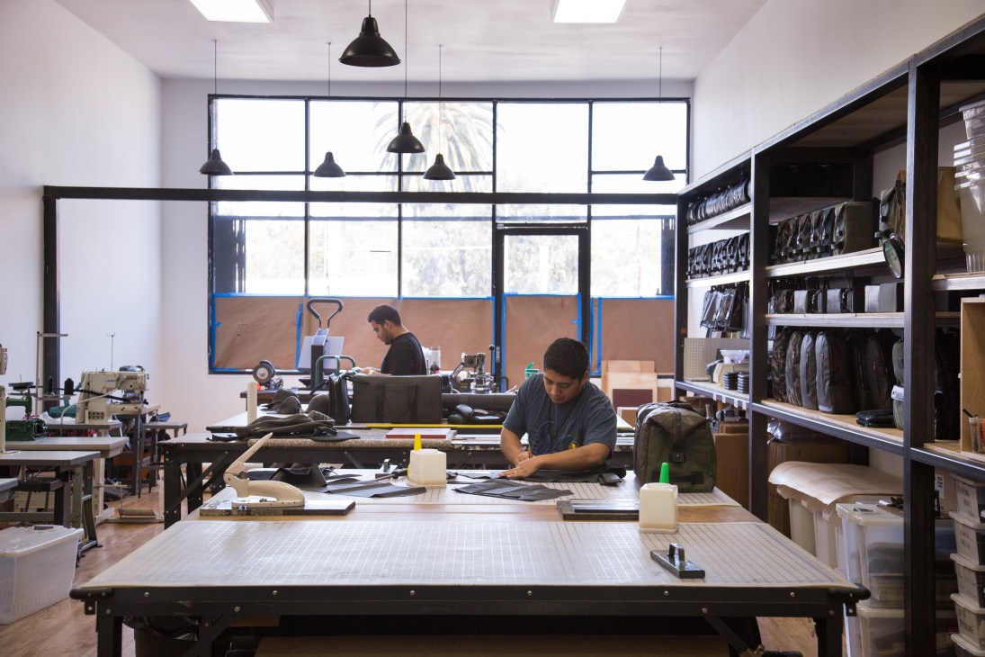 Workers in a bright, open-plan workshop with large windows, workbenches, and organized shelving displaying leather goods and materials.