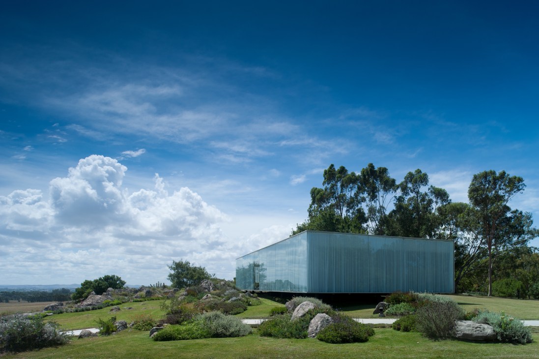 Modern glass and metal structure nestled among boulders and grassland with eucalyptus trees under blue sky.