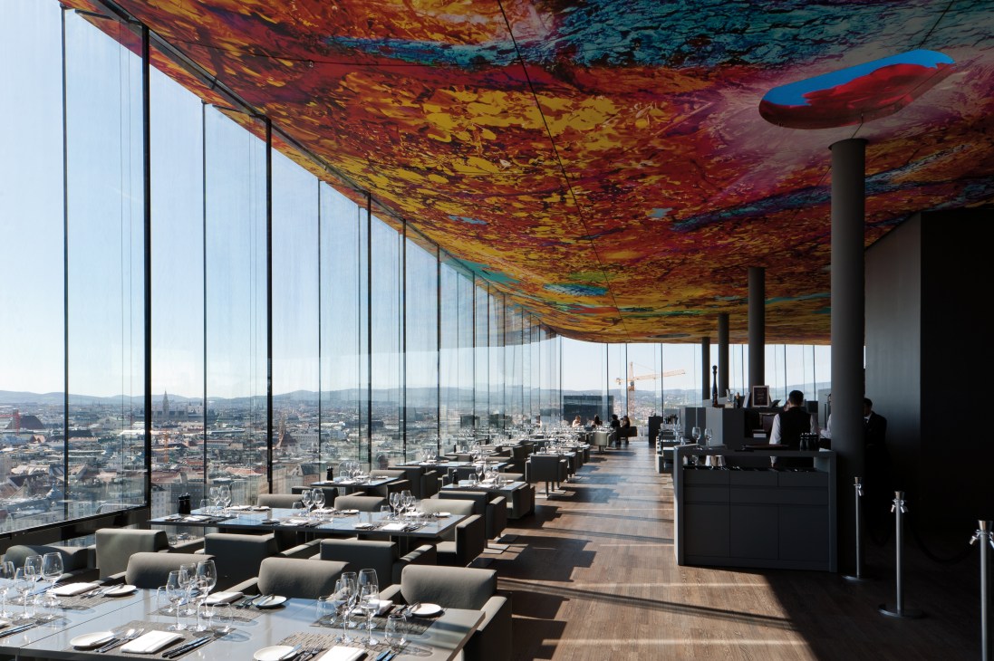 Le Loft restaurant interior with floor-to-ceiling windows overlooking Vienna, colorful ceiling artwork, and dining tables.