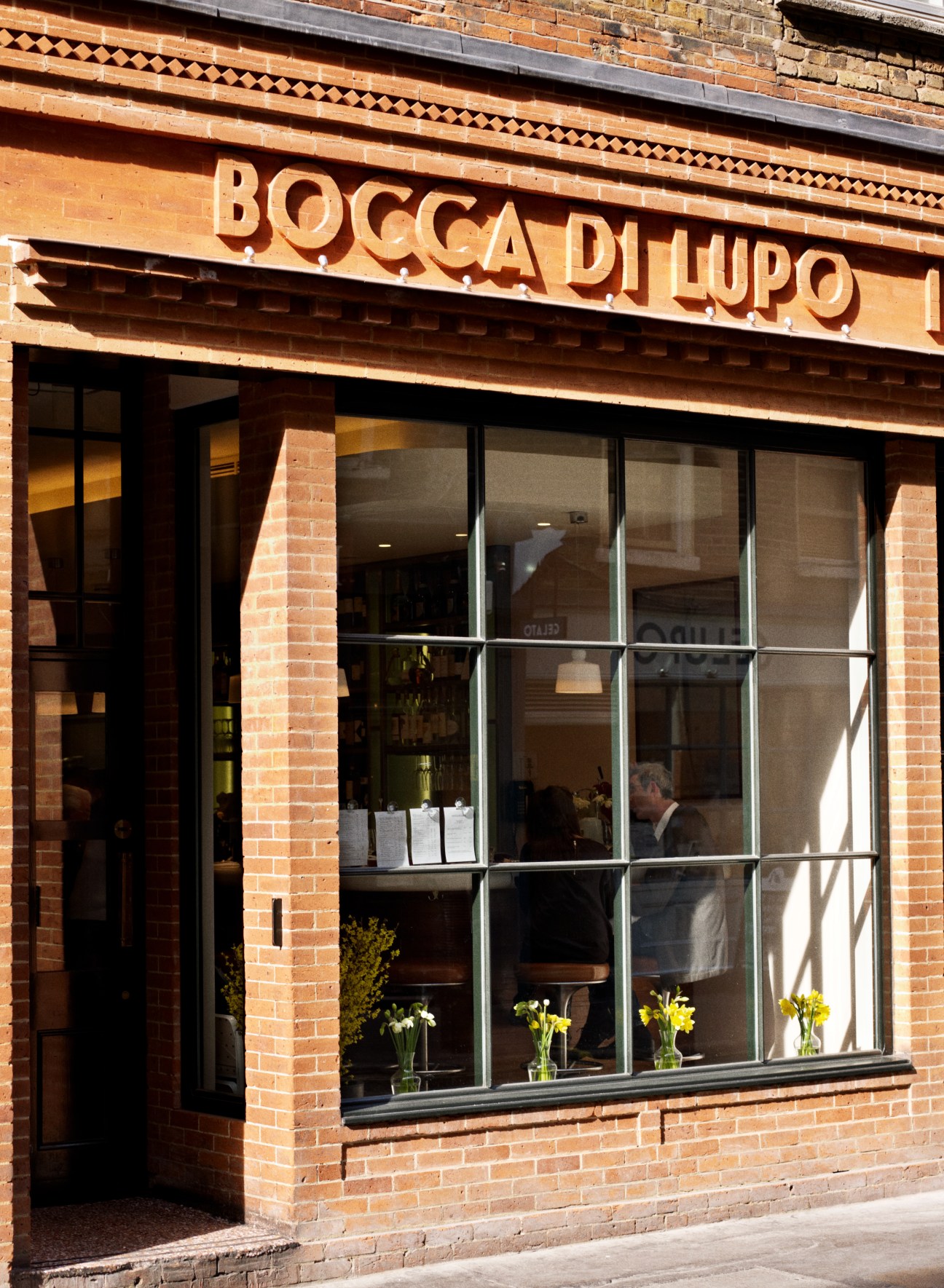 Bocca di Lupo restaurant storefront in Soho with brick facade and large windows displaying yellow flowers.