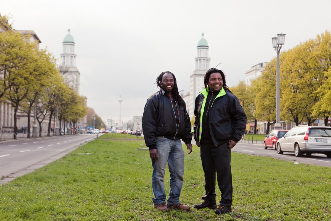 Two men standing on a grassy area in Berlin with classical buildings and tree-lined avenue in background