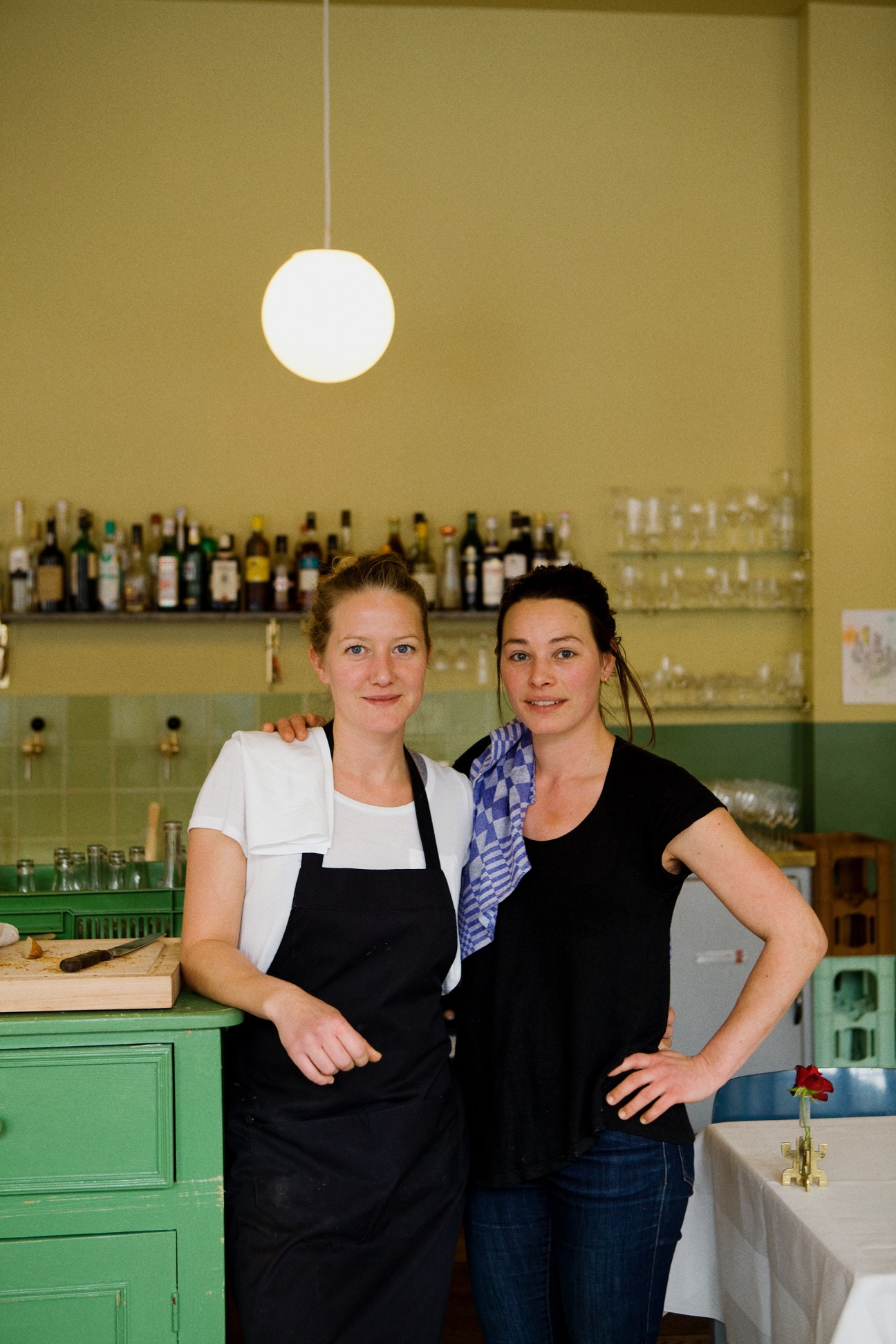 Two women standing together in a bar with yellow walls, green cabinetry, and wine bottles on shelves behind them.