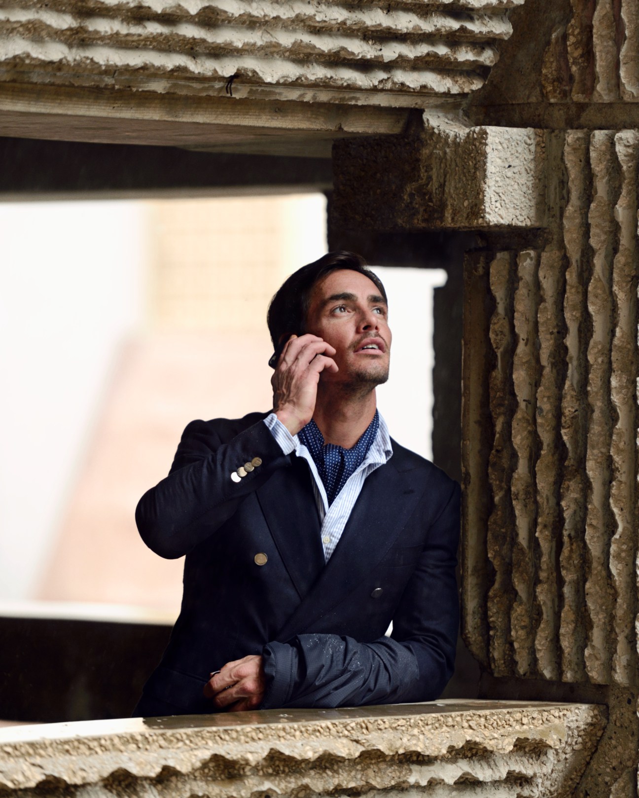 Man in Tomorrowland jacket and Dunhill coat talking on phone against weathered brick wall.