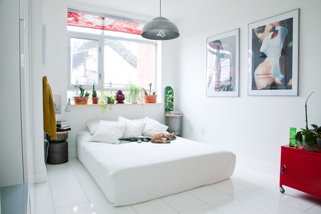 A bright, minimalist bedroom with a white bed, potted plants on the windowsill, and framed artworks on the walls.