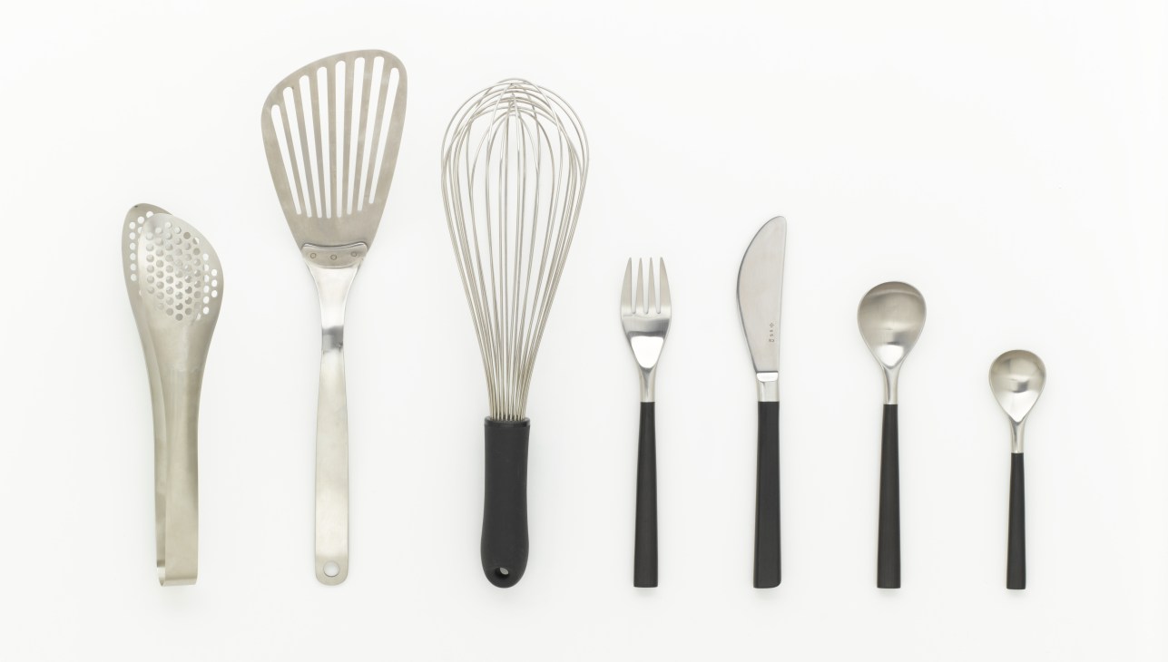 Sori Yanagi's handcrafted steel kitchen utensils including skimmer, slotted spoon, whisk, fork, knife, and spoons with black handles.