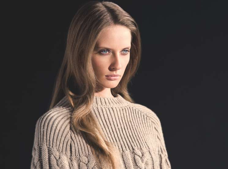 Model wearing a Louis Vuitton cape over a cream cable-knit sweater against a dark background.