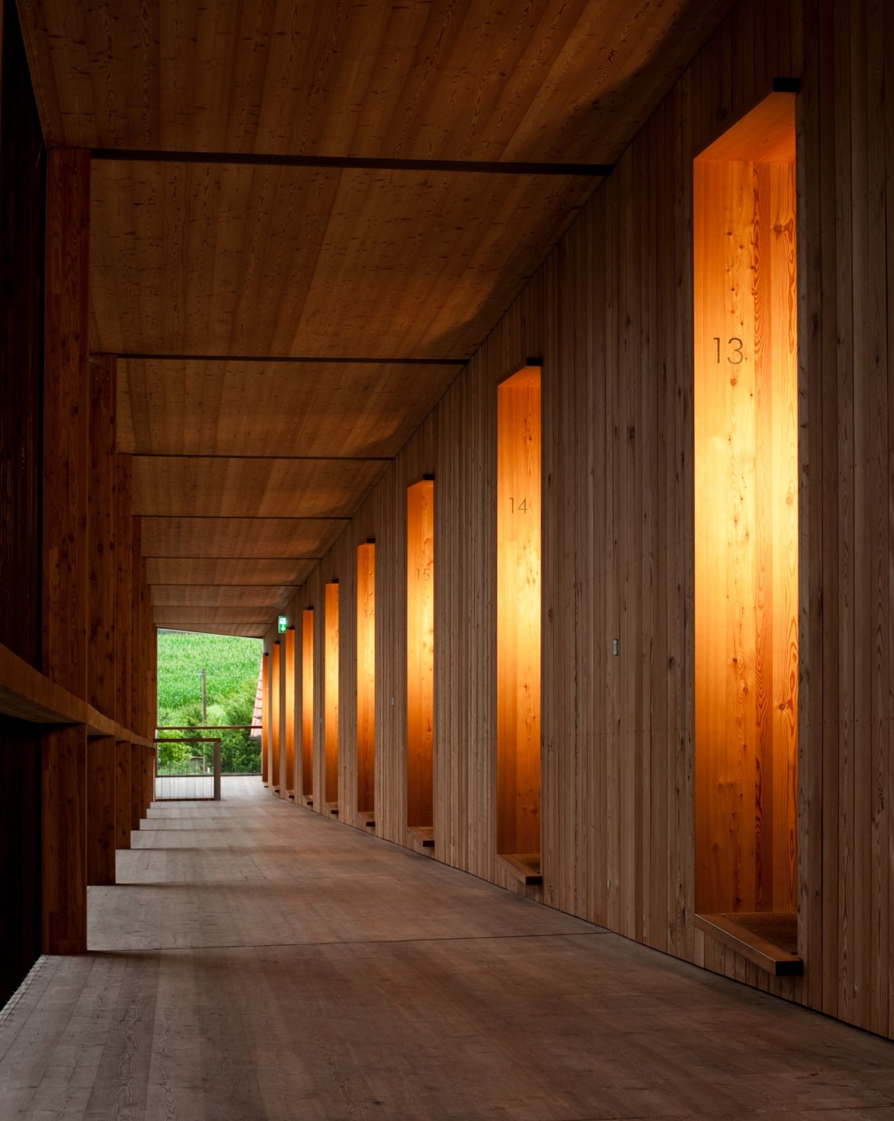 Interior corridor of Hörger Biohotel with wooden walls, illuminated orange room dividers numbered 13-15, and green orchard visible through the entrance.