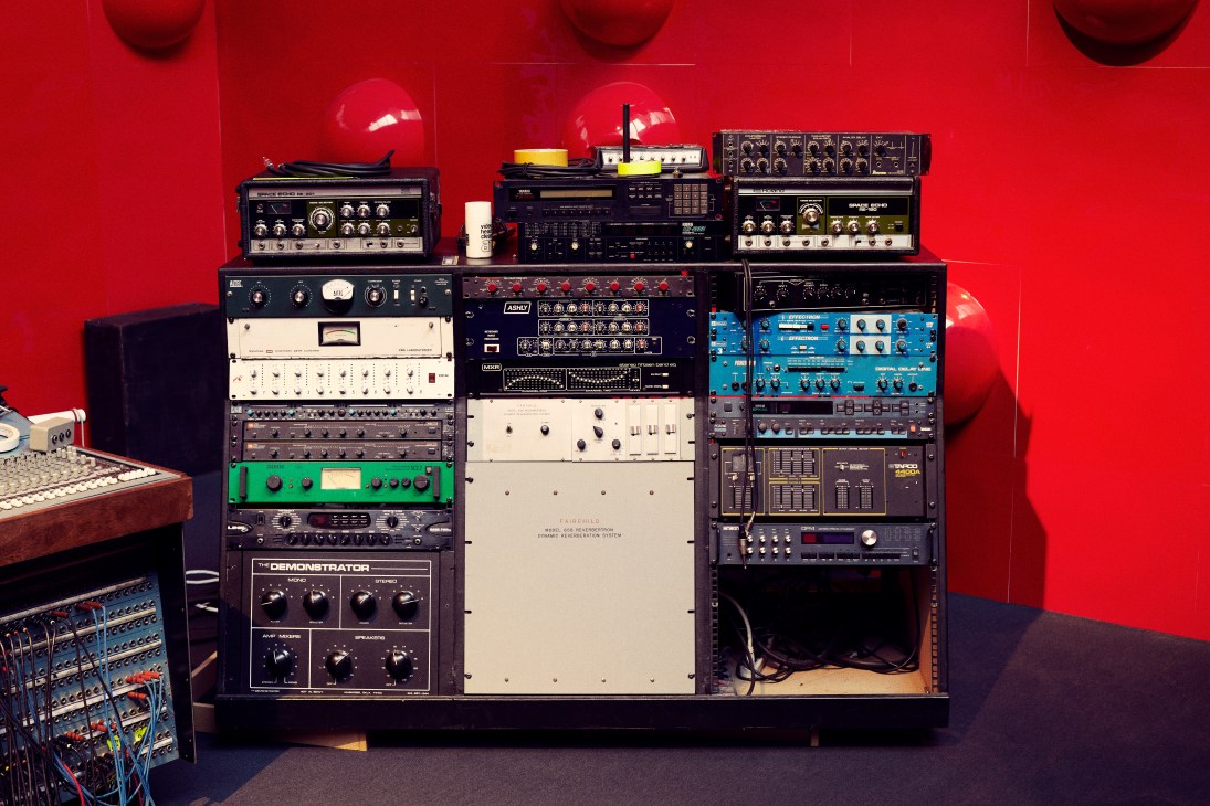 Wall of vintage synthesizers and recording equipment stacked against a vibrant red studio wall.