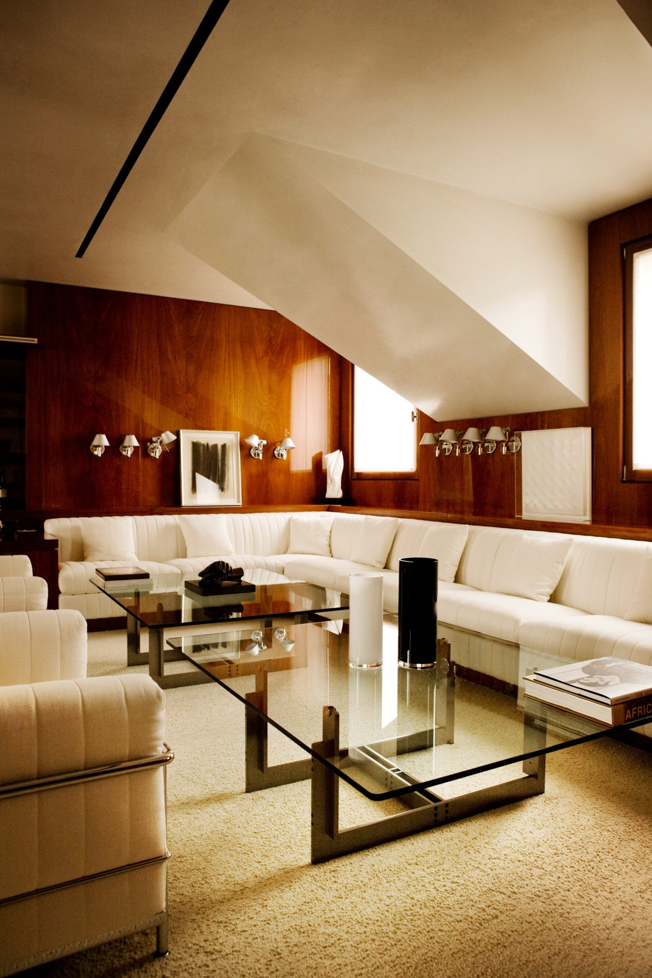 Modern living room with cream upholstered seating, mahogany wood panelling, and glass coffee tables under sloped ceiling.
