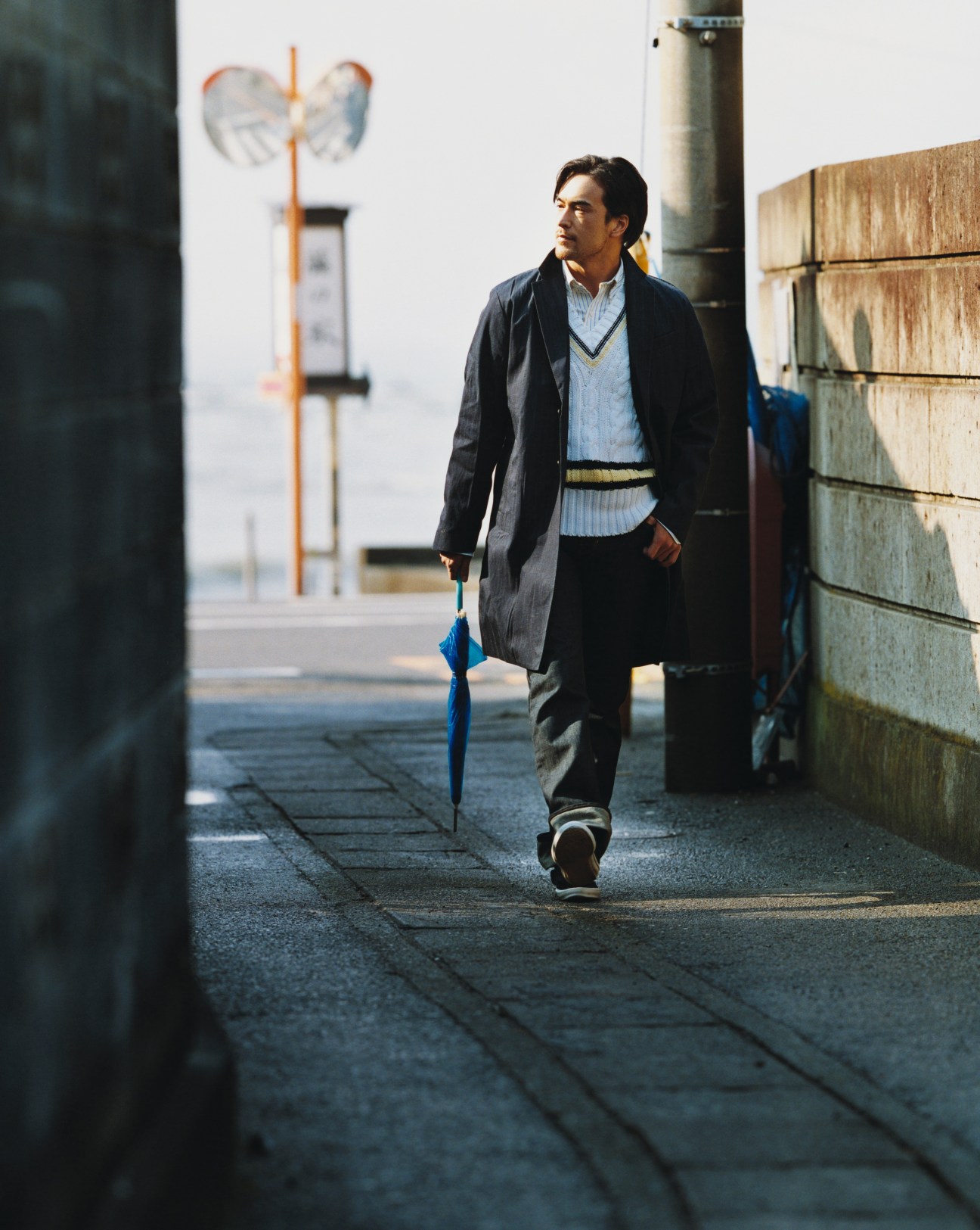 Man wearing Prada denim coat and Black Fleece by Brooks Brothers jumper walks along Hayama waterfront holding blue umbrella.