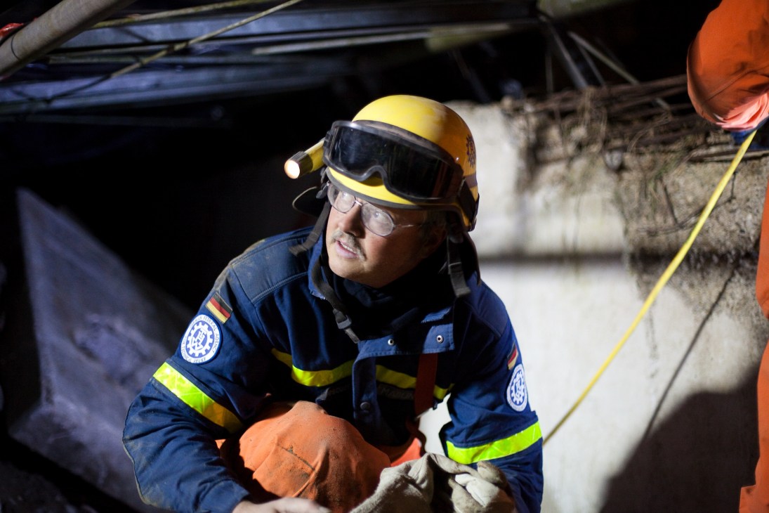 THW volunteer emerges from a tunnel wearing yellow helmet, safety goggles and blue uniform with reflective strips during rescue training.