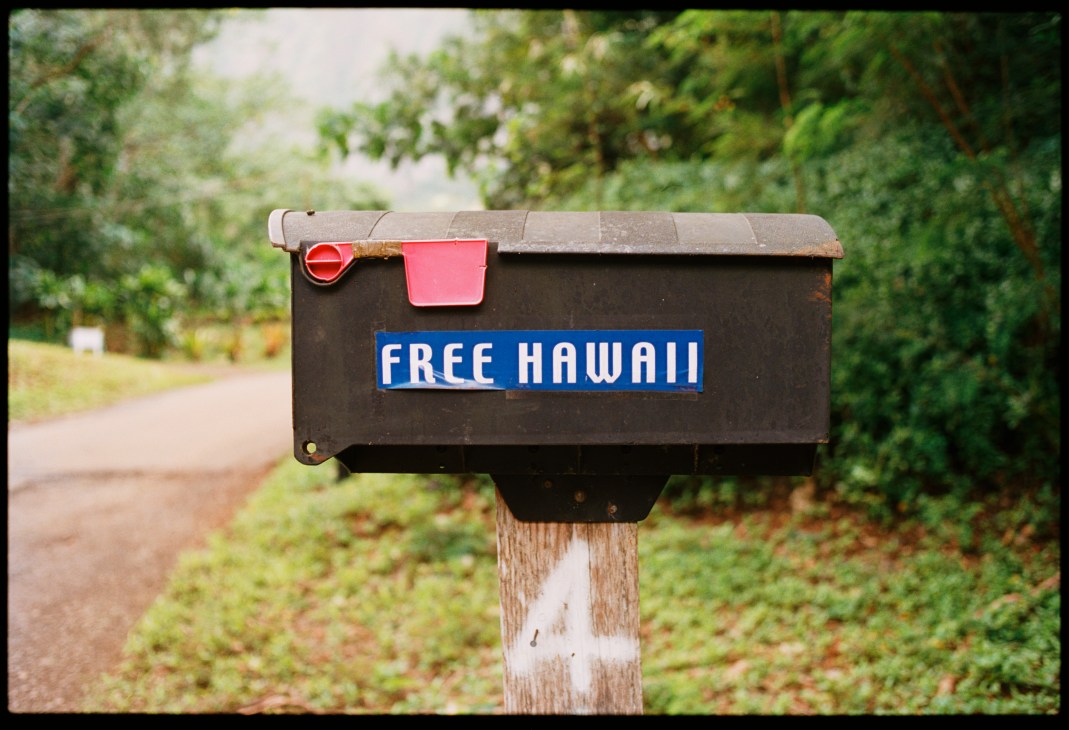 A black mailbox with "FREE HAWAII" text and a red flag mounted on a wooden post surrounded by green vegetation.