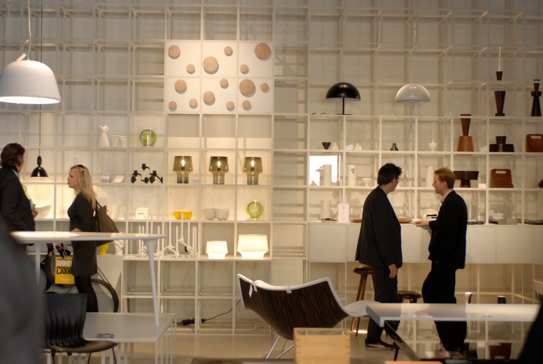 Modern design showroom with white grid wall display shelving featuring lighting fixtures and decorative objects, visitors conversing in foreground.