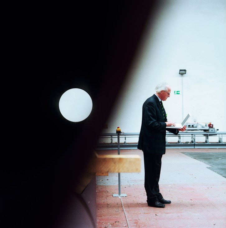 Dietrich Stein, an older man with grey hair wearing a dark suit, stands at a test track holding a laptop computer while controlling a CargoCap model vehicle.