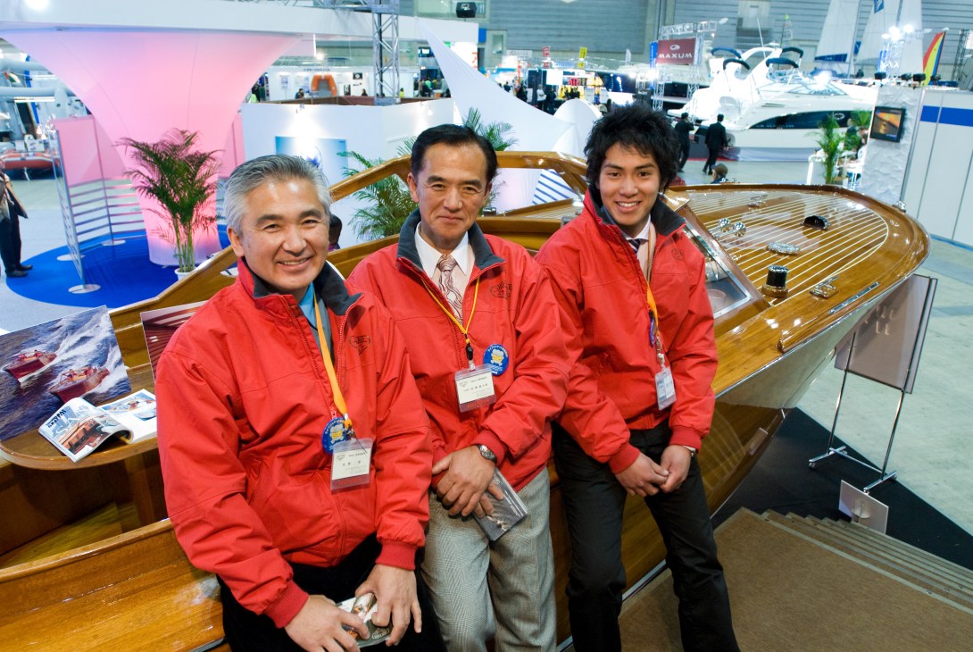 Three men in red jackets at a boat show stand with a wooden boat behind them.