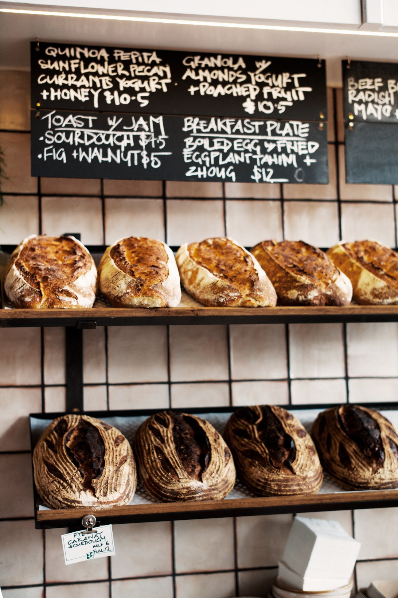 Artisan breads displayed on wooden shelves against white subway tiles with a handwritten menu board above.