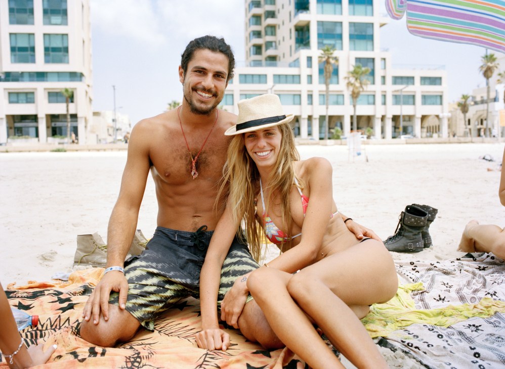Two young people sitting on the sand at Banana Beach in Tel Aviv with modern high-rise buildings behind them.