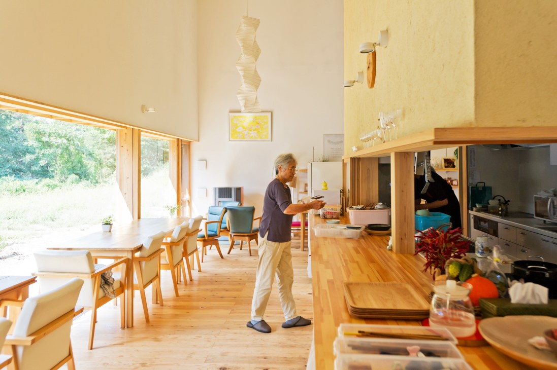 An elderly man in a brown shirt stands in a bright, modern restaurant interior with wooden furnishings and large windows overlooking green landscape.