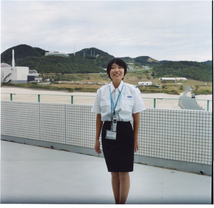 JAXA PR staff member Mis Yuji in white shirt and JAXA badge standing at Tanegashima Space Center with launch facilities visible behind her.