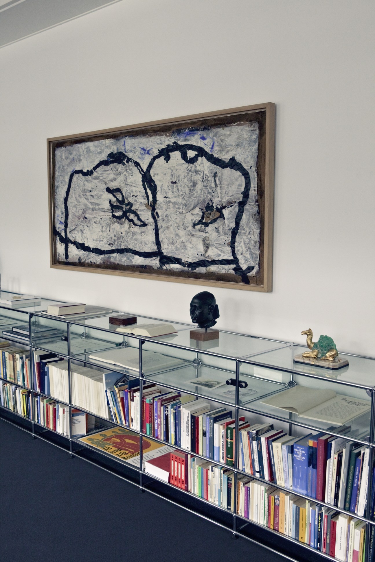 Abstract artwork with dark blue lines on textured white canvas displayed above a glass library shelf with books and sculptures.
