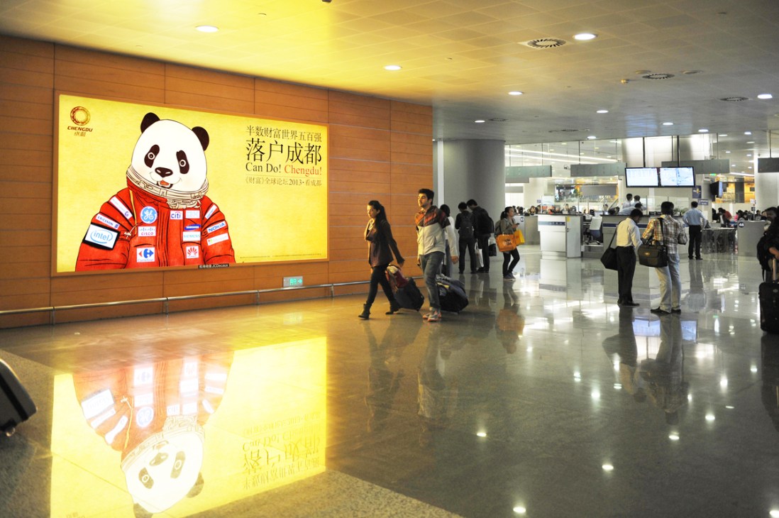 Giant panda mascot wearing red tracksuit on airport terminal billboard advertising Chengdu's Fortune 500 forum.