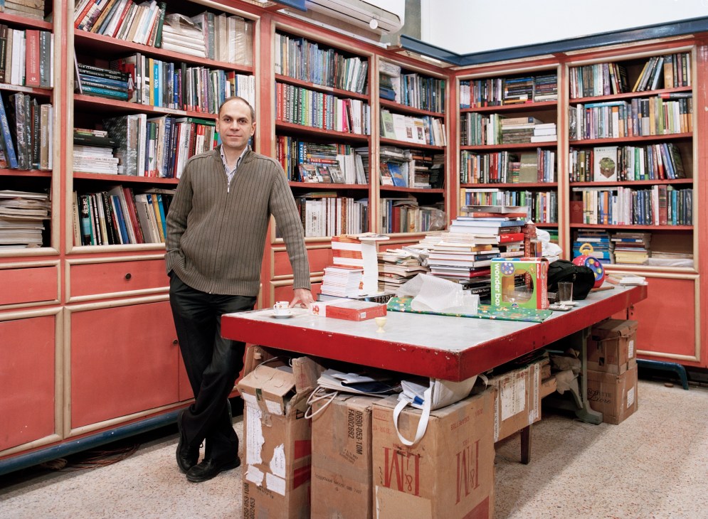 Man in pinstriped sweater standing at red desk in bookstore with red shelving and books.