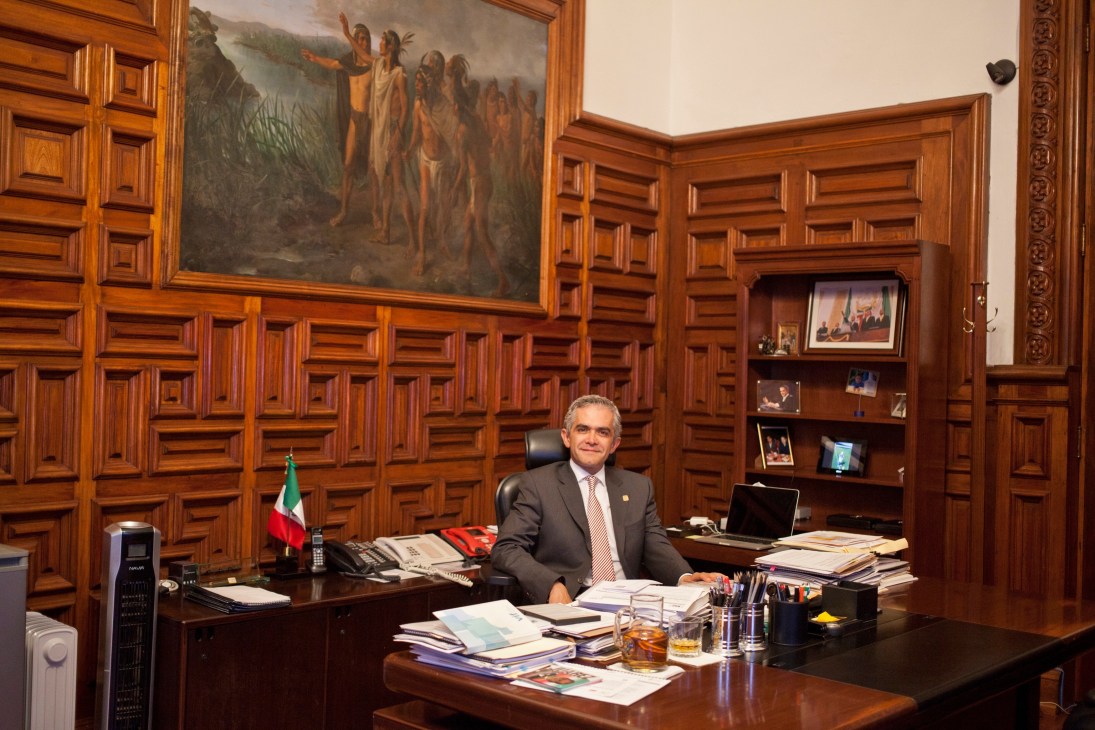 Mayor Miguel Ángel Mancera seated at his desk in an ornate wood-paneled office with a Mexican flag.