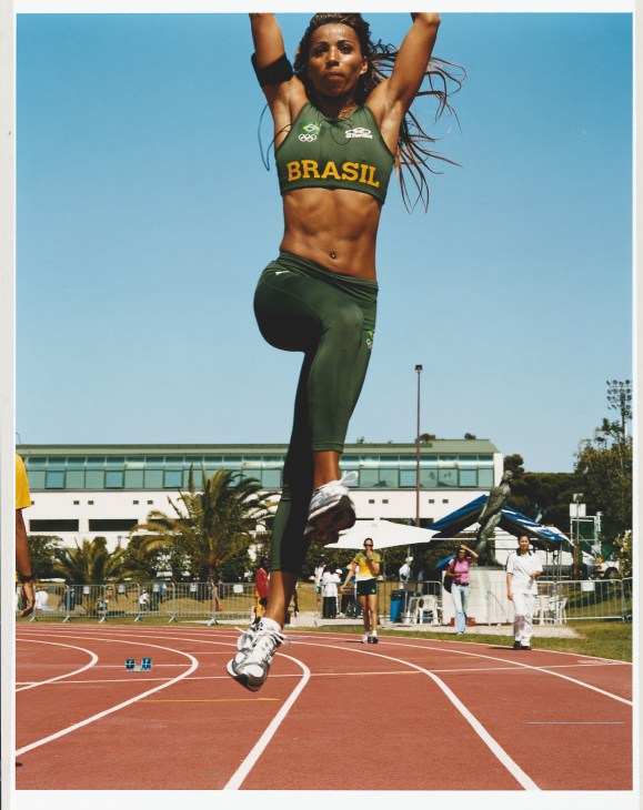 Brazilian athlete in green Brasil tracksuit performing long jump warm-up on red track.