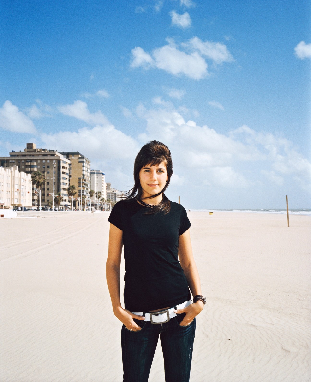 Young woman standing on a beach in Cádiz with apartment buildings and palm trees behind her.