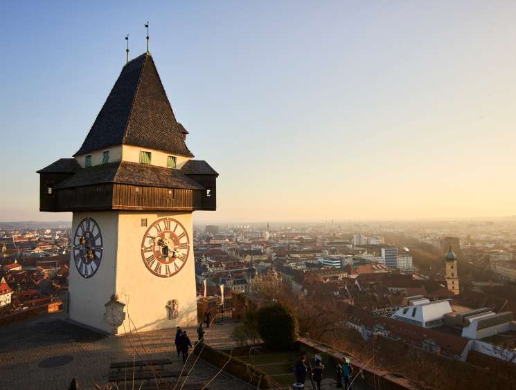Clock tower with wooden gallery and pointed roof overlooking red-roofed cityscape at sunset