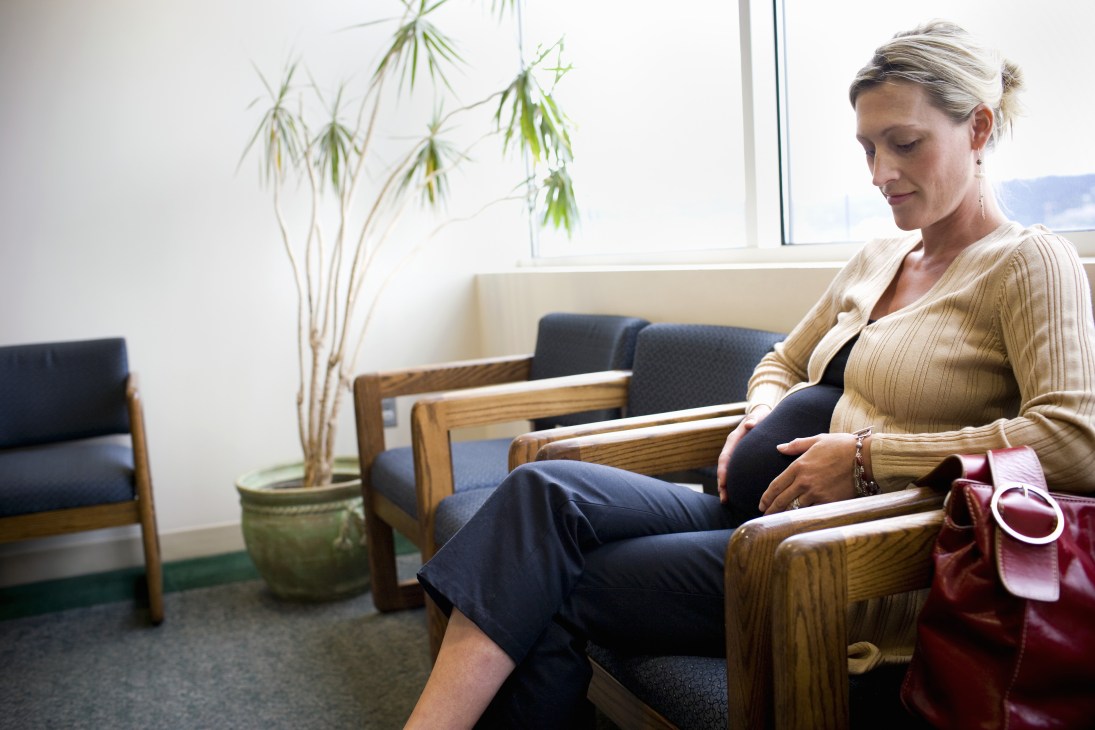 Pregnant woman sitting in a waiting room chair with a potted plant nearby.