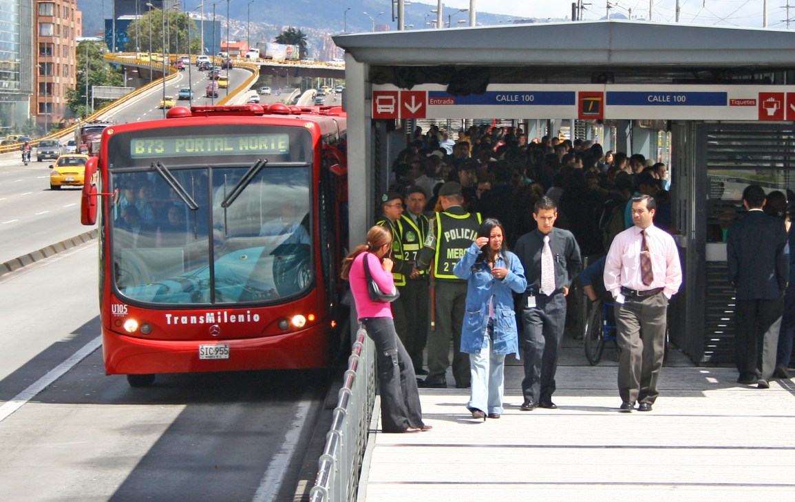 Red Transmilenio bus at a station stop in Bogotá with passengers boarding and police officers present.