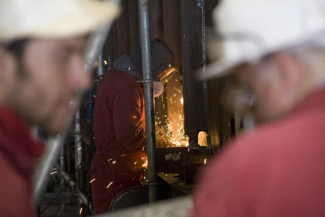 A welder in protective red clothing performs welding work, with bright orange sparks flying from the joint.