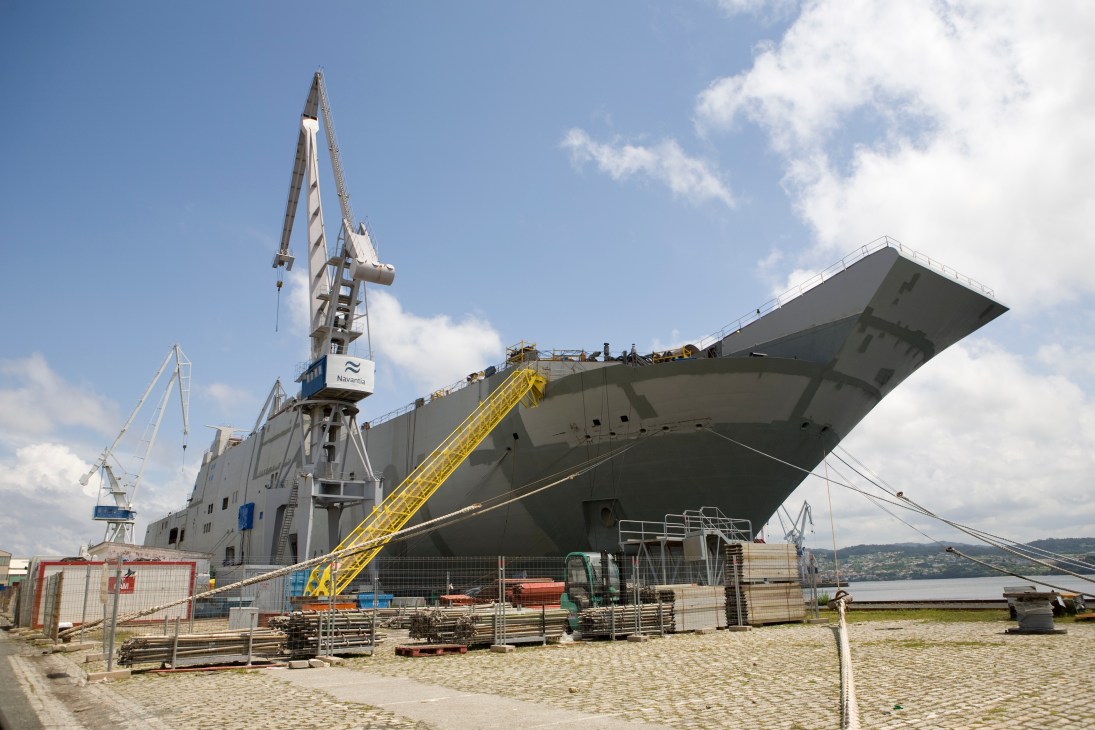 The Juan Carlos I, a large grey landing helicopter dock ship under construction at Navantia shipyard with cranes and equipment nearby.