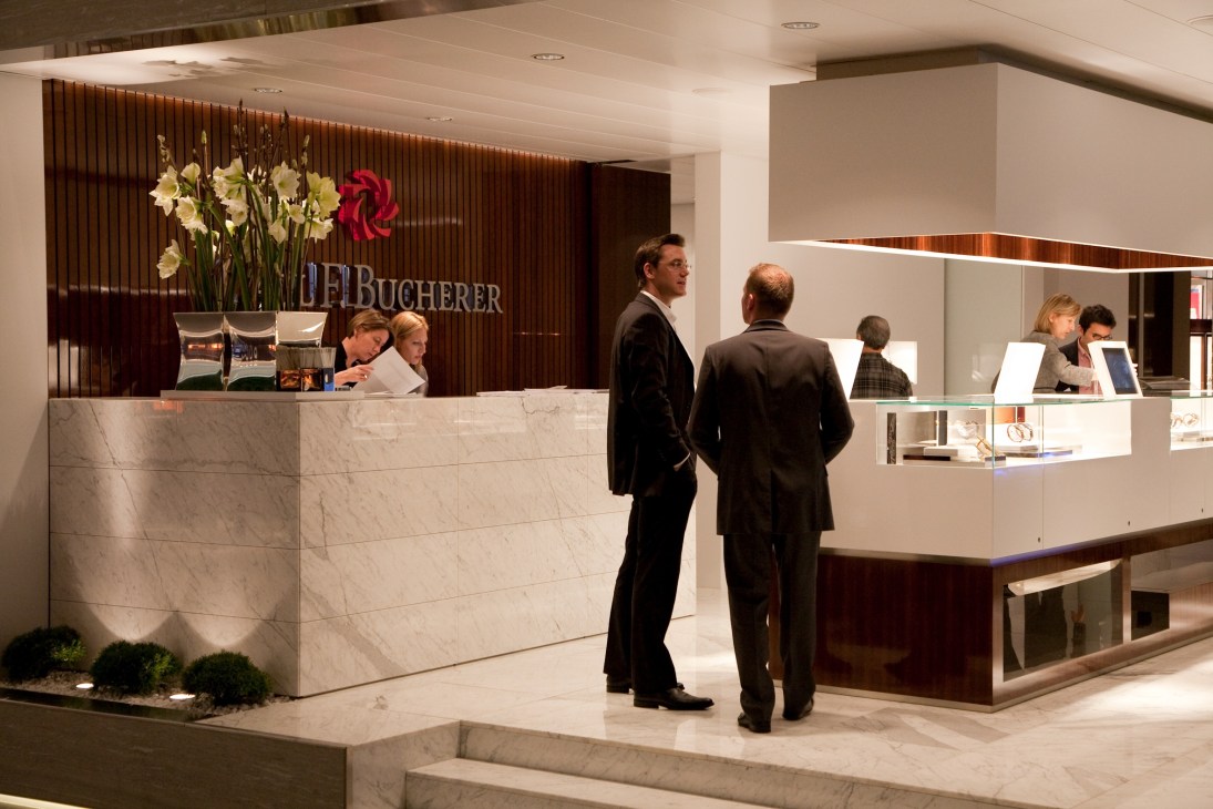 Carl F Bucherer's exhibition stand at a watch fair with marble counter, wooden backdrop, and visitors in business attire.