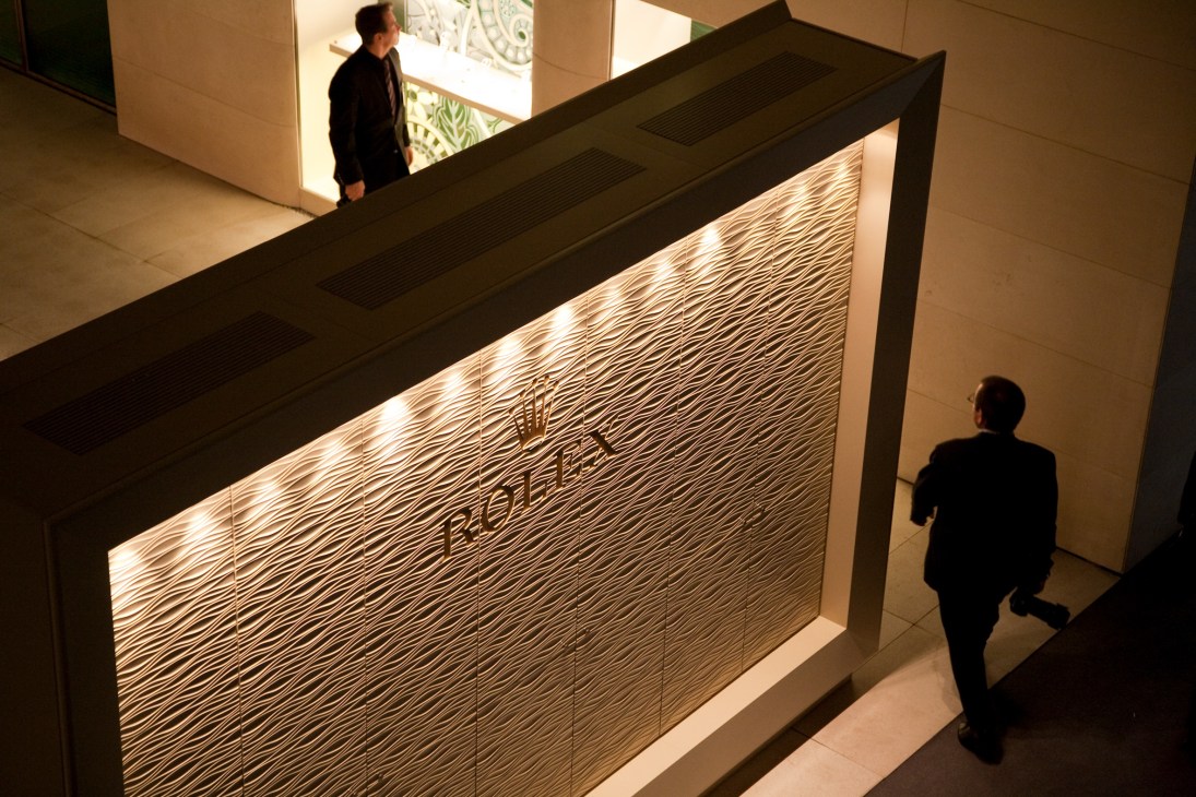Rolex's illuminated exhibition stand with textured wall panel and logo, visitors walking nearby at watch fair.
