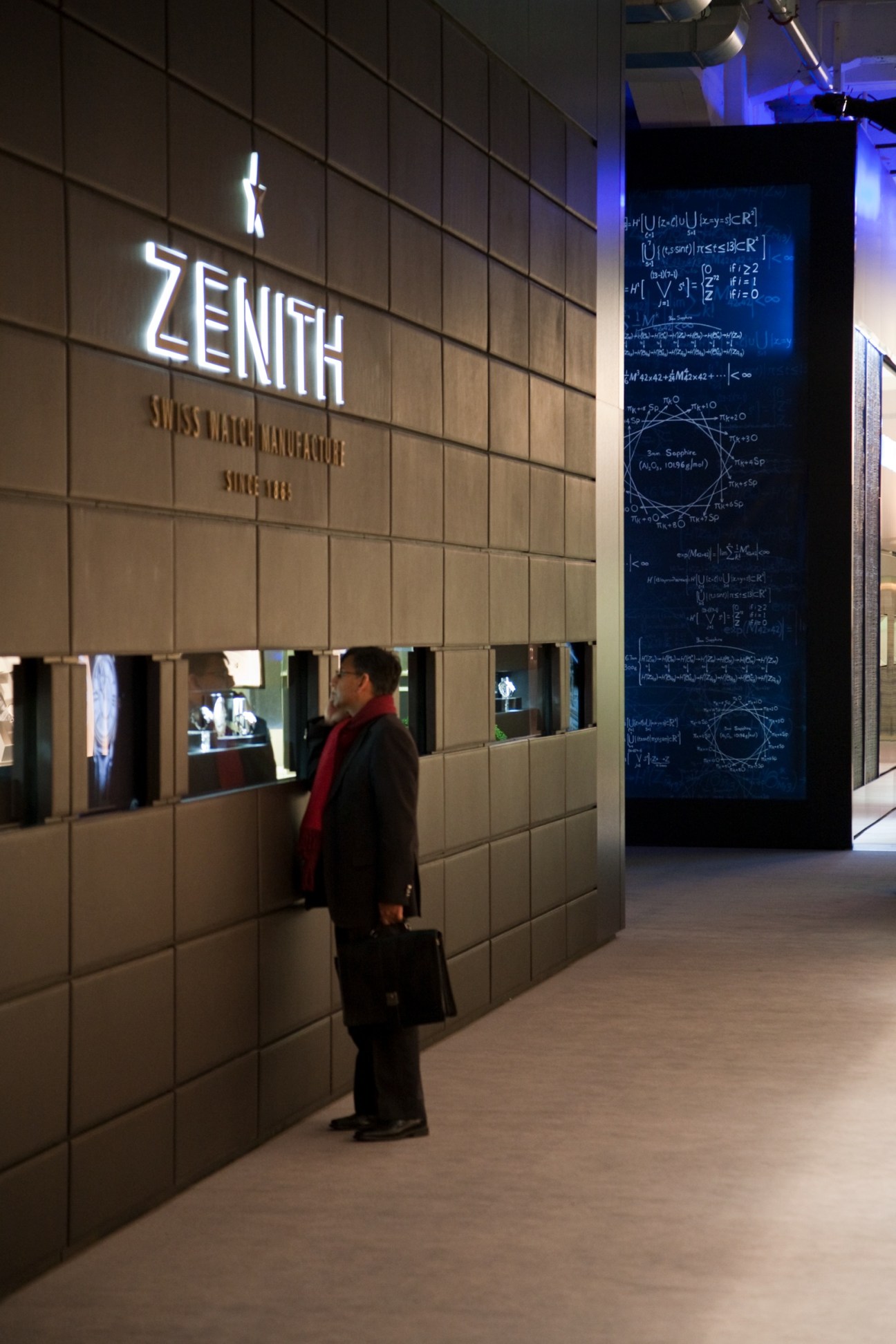 Zenith exhibition window at watch fair with illuminated signage and mathematical equations on display screen.