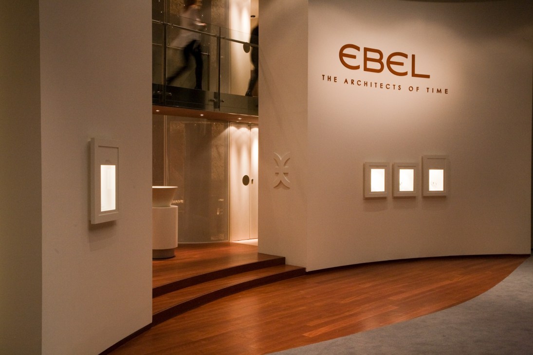 Ebel's exhibition stand at Basel watch fair with minimalist white walls, wooden flooring, and illuminated display cases.