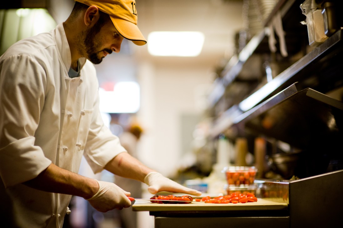 Chef in white chef's coat and yellow cap preparing food in a professional kitchen.