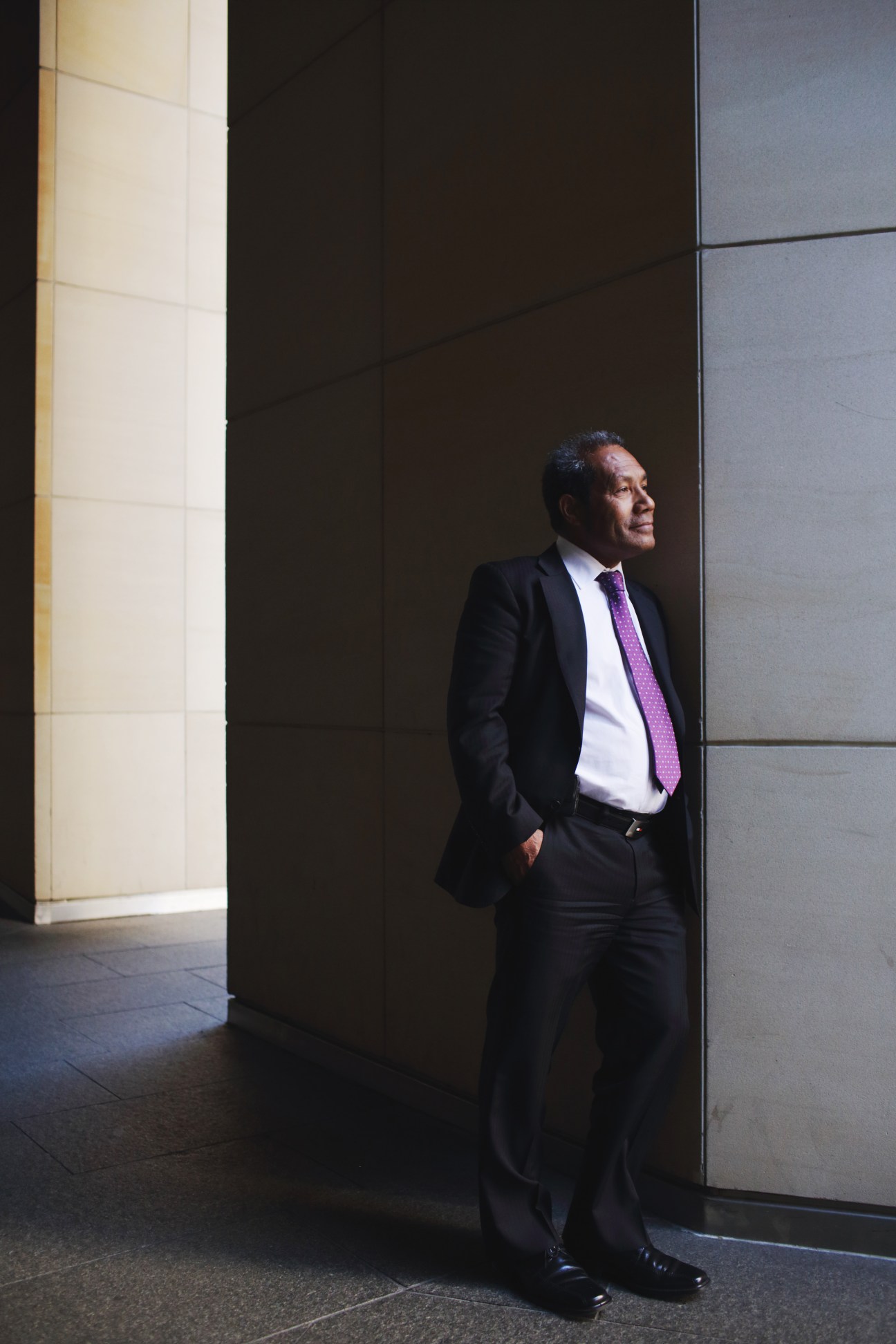 Man in dark suit and purple tie standing in modern corridor with geometric wall panels.