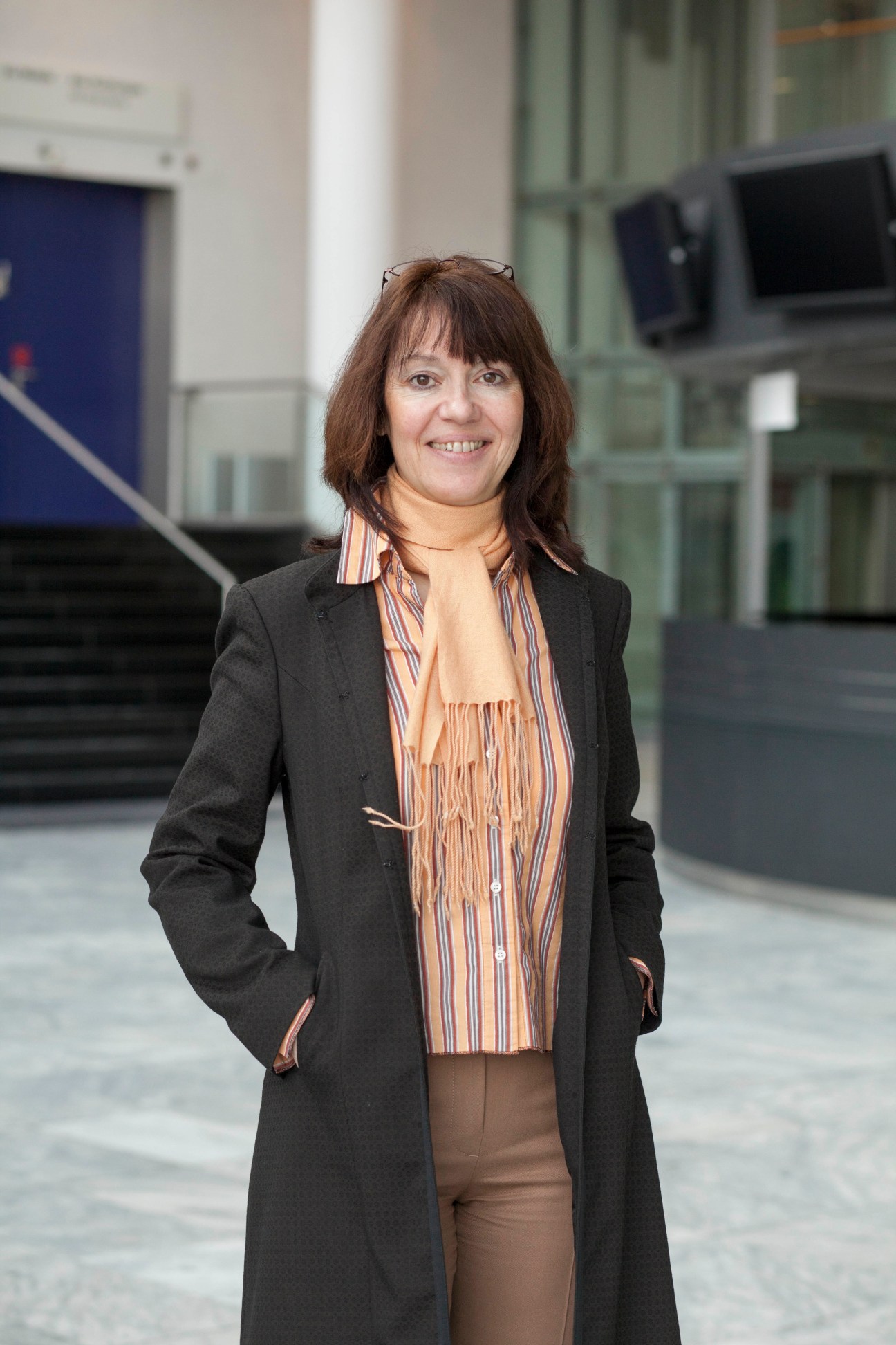 Woman in black coat and striped shirt standing in modern exhibition hall corridor.